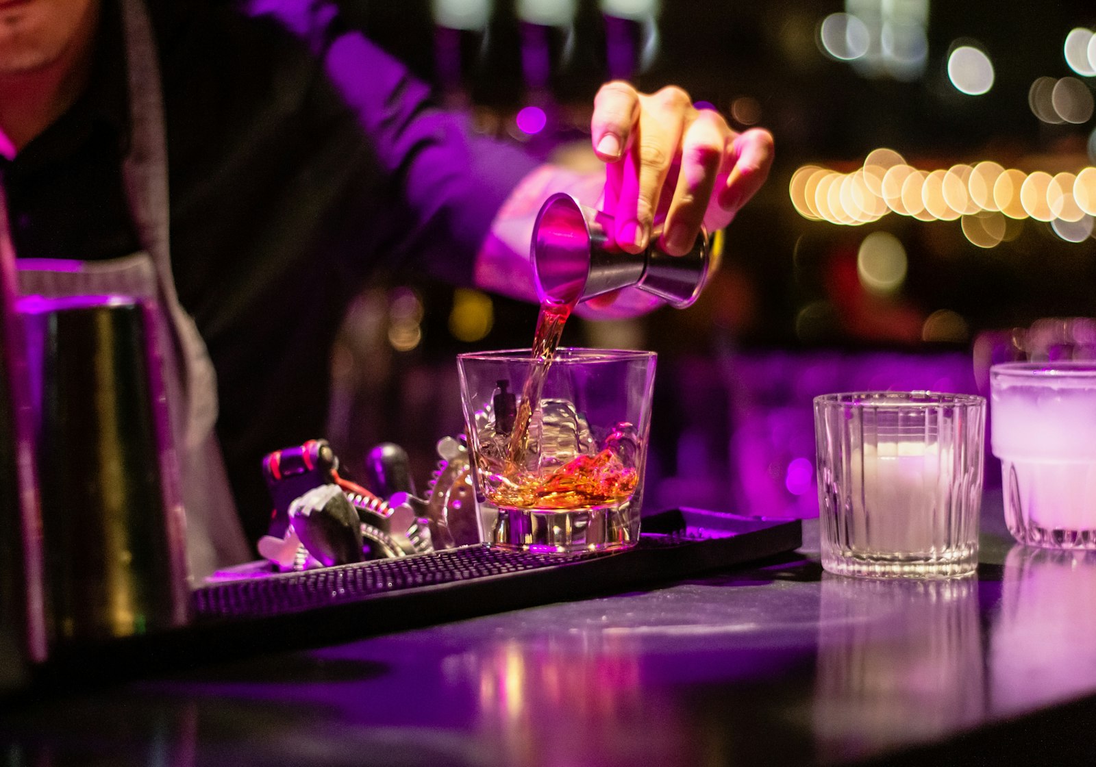 Bartender pouring whiskey from a jigger into a rocks glass under purple light